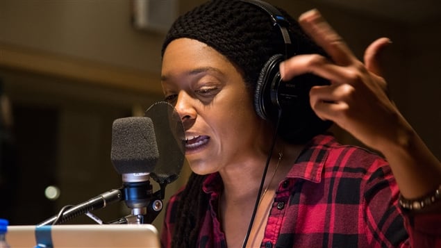 A woman in a red flannel shirt and black cap speaks into a microphone in a radio studio while gesturing with one hand