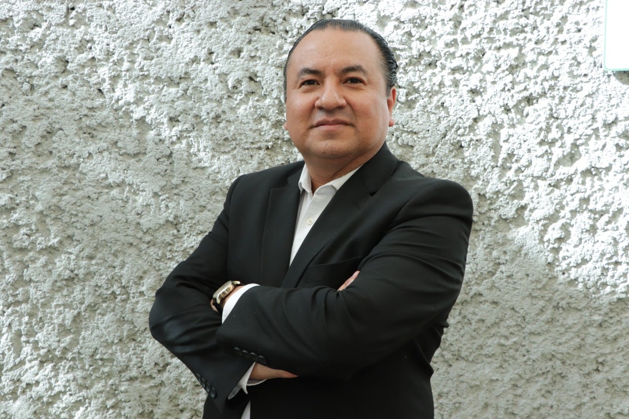 A man in a suit stands with his arms crossed next to a wall.