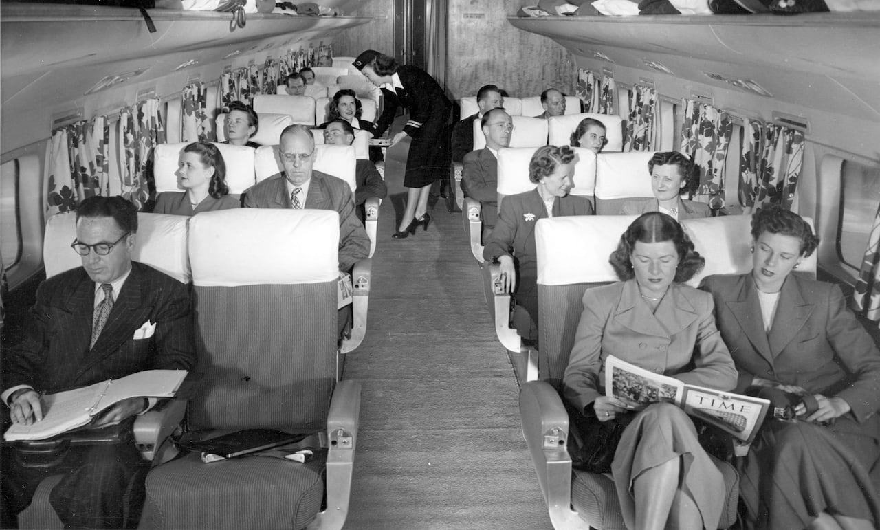 Old black and white photo of men and women sitting in their seats on an airplane. They are dressed formally.