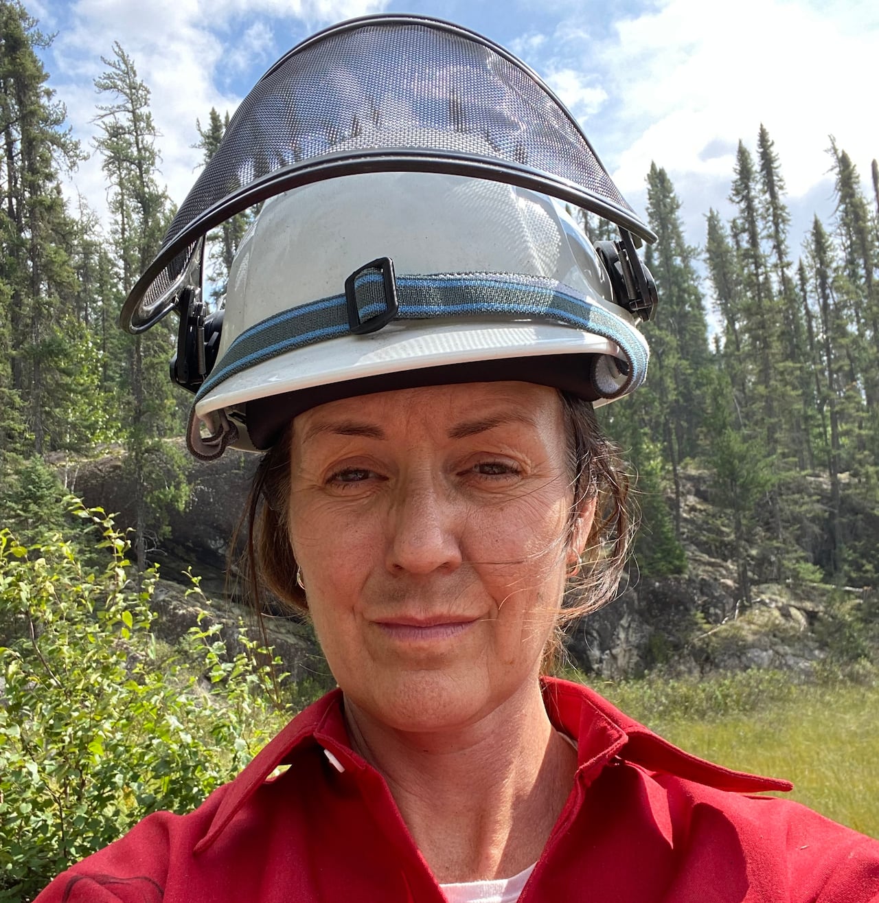 A woman in a forest wearing a firefighters helmet and red uniform.