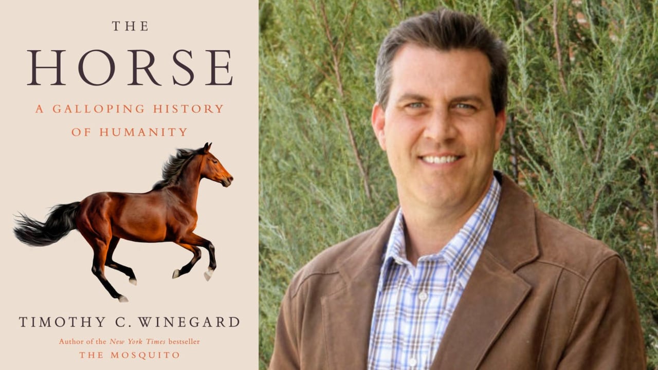 A man with dark hair brushed back is wearing a brown suede jacket and white/blue plaid dress shirt. He is smiling and standing in front of a large pine bush.