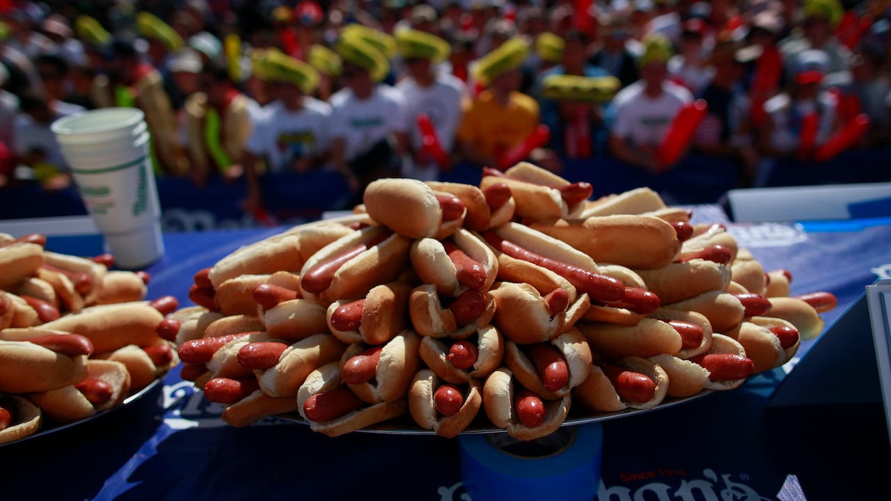 A large plate piled with hot dogs rests on a table with a blue counter top. A large crowd can be seen in the background.