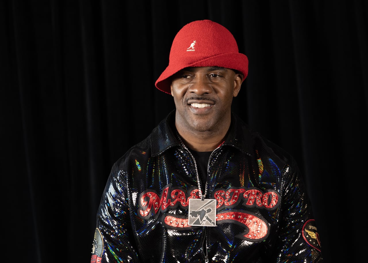 Portrait of a man smiling as he looks to one side, wearing a Kangol-brand red bucket hat and a jacket with the word "Maestro" emblazoned across the front in red, stylized, rhinestone-covered letters. 