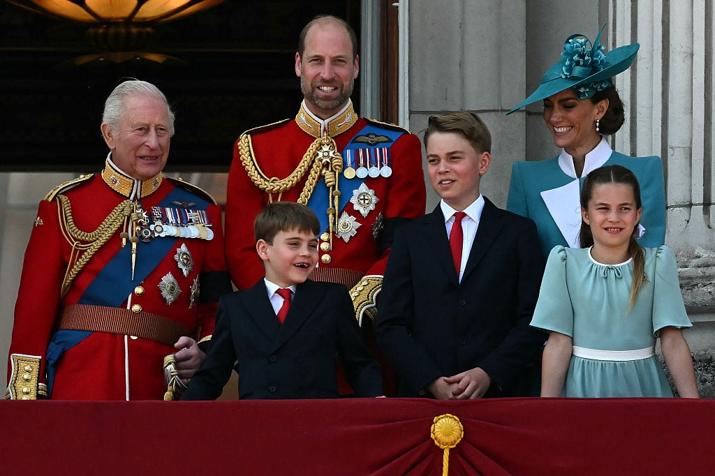 Three adults and three children stand on a balcony of a large stone building with an open entryway behind them.