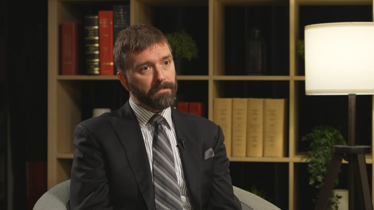 A man in a suit sitting in a chair in front of a bookshelf.