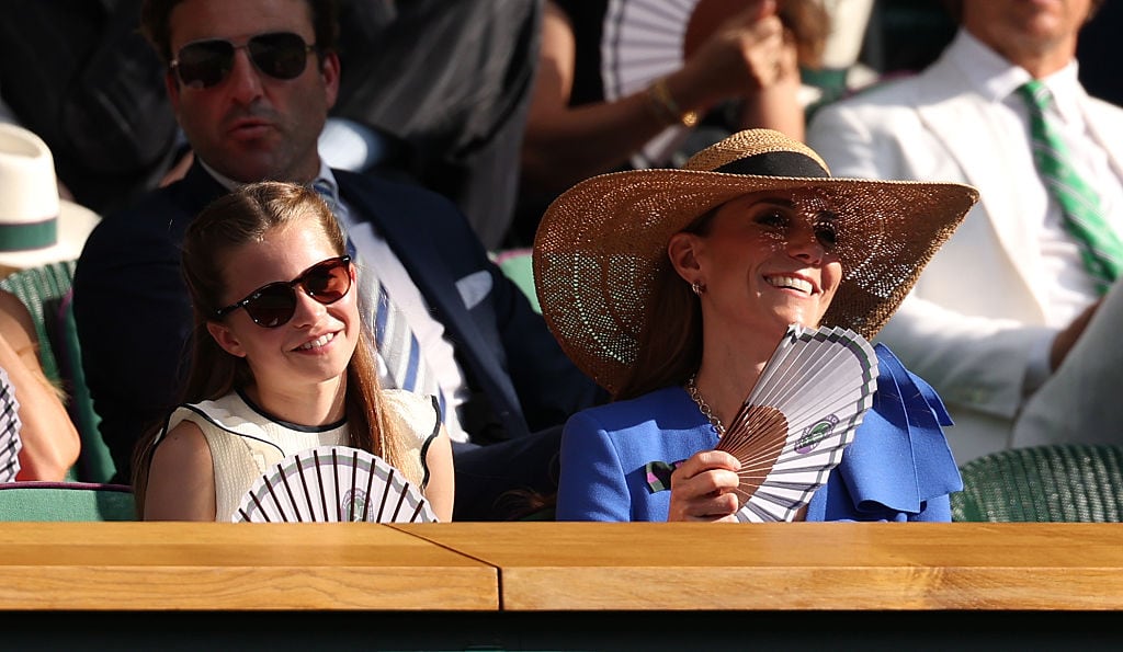 Two people seated beside one another wearing sunglasses hold fans in front of their faces.