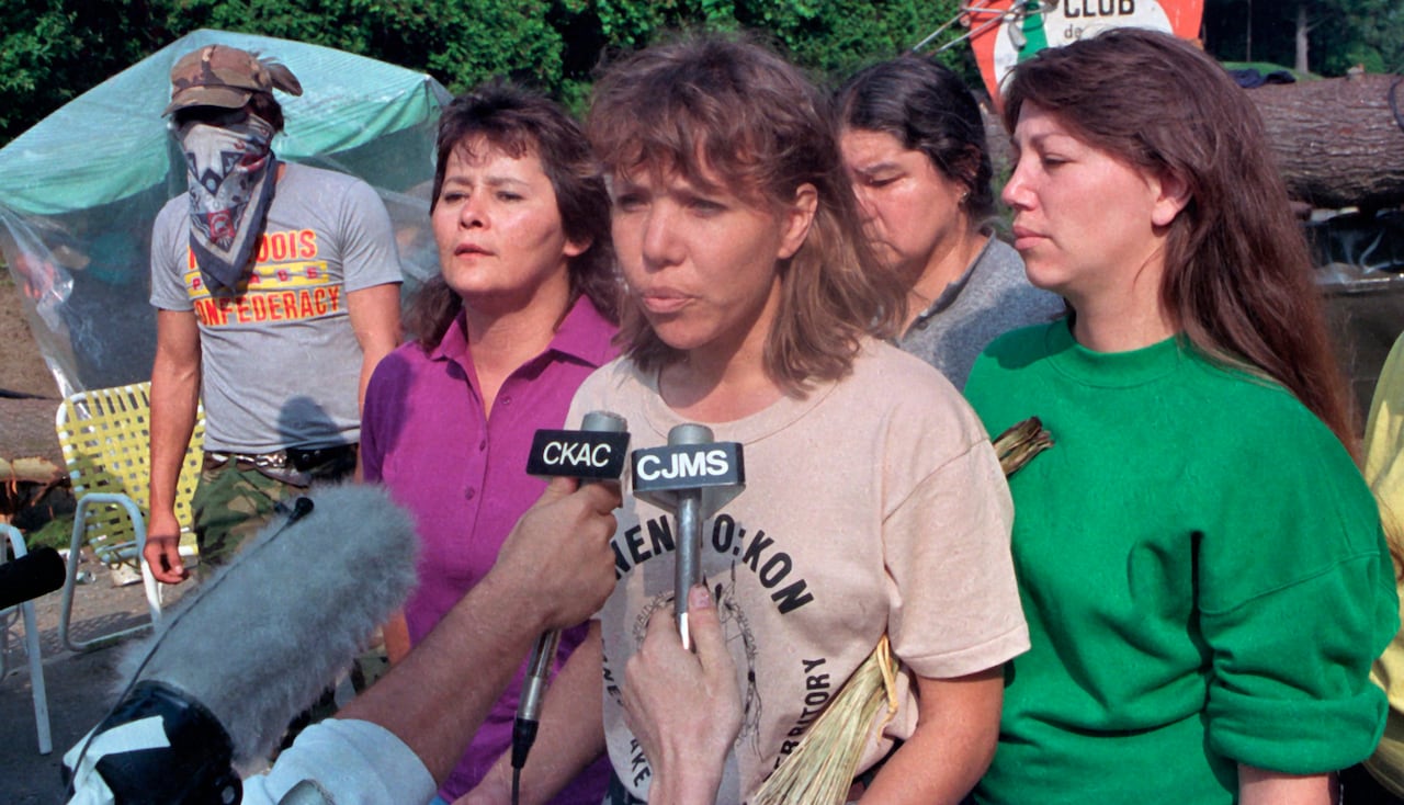 A woman talks into microphones flanked by supporters with a blockade in the backgroun.