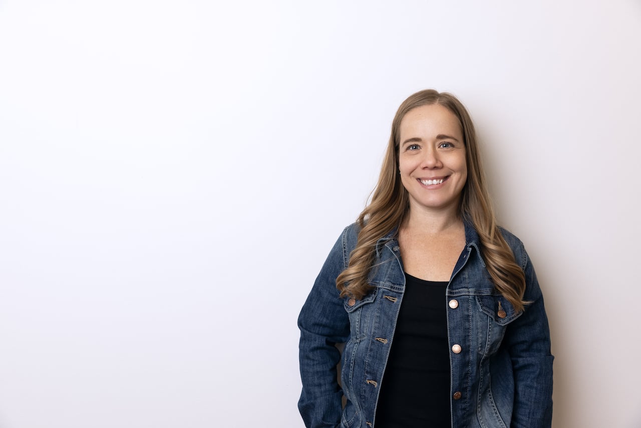 A woman in a jean jacket stands smiling against a wall