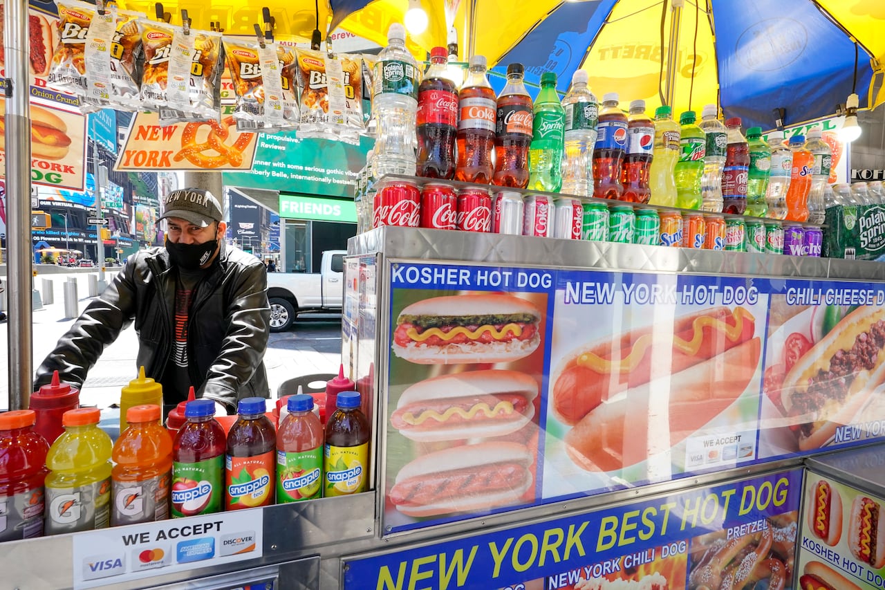 A man wearing a jacket and face mask stands at his hot dog cart, lined with images of hot dogs and covered in drink bottles and soda cans.