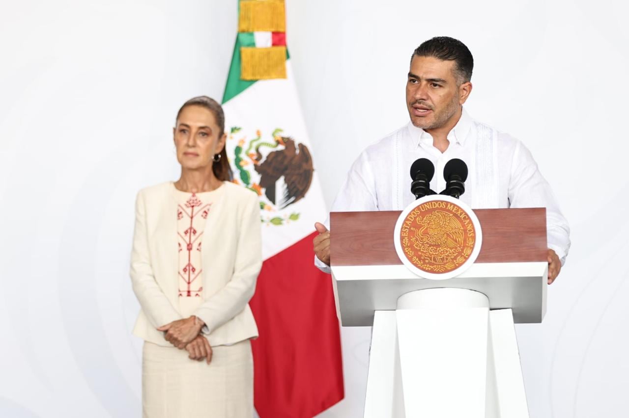 A man in a white shirt stands a podium while a woman in the background looks on.