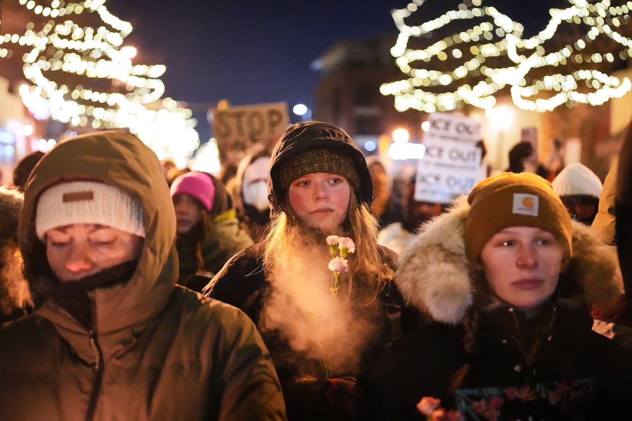 Three women are seen in a crowd at night lit by candles and holiday lights behind them.