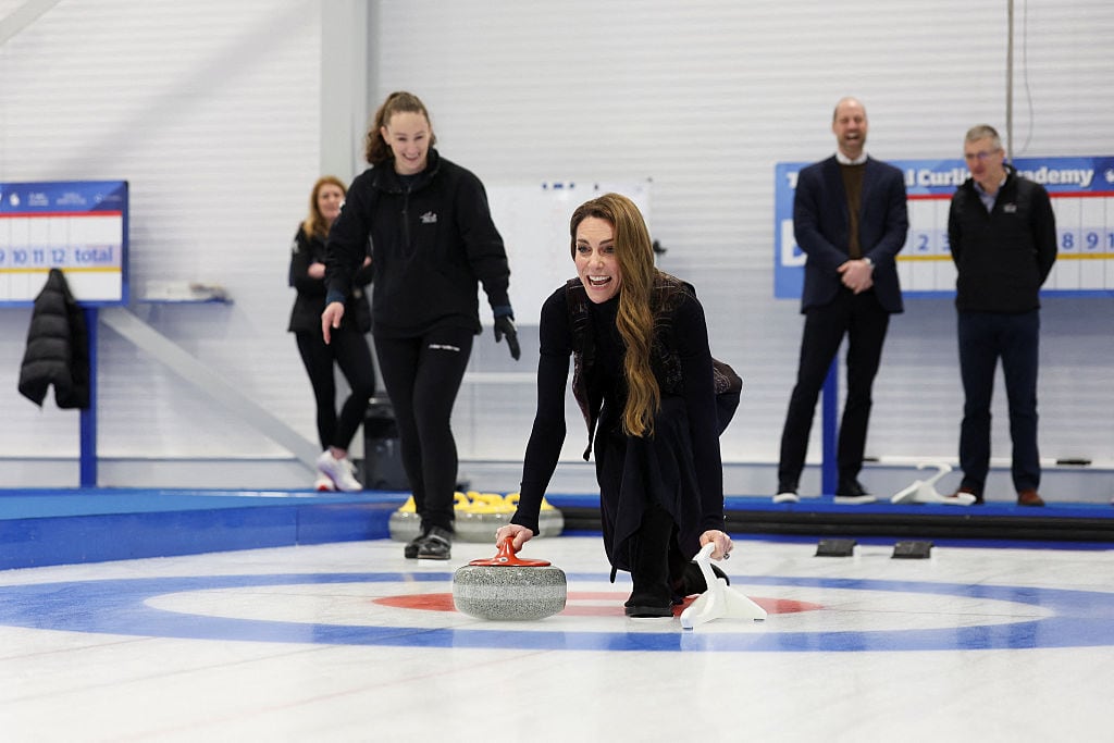 A person kneels on ice to release a curling rock as several people standing nearby smile.