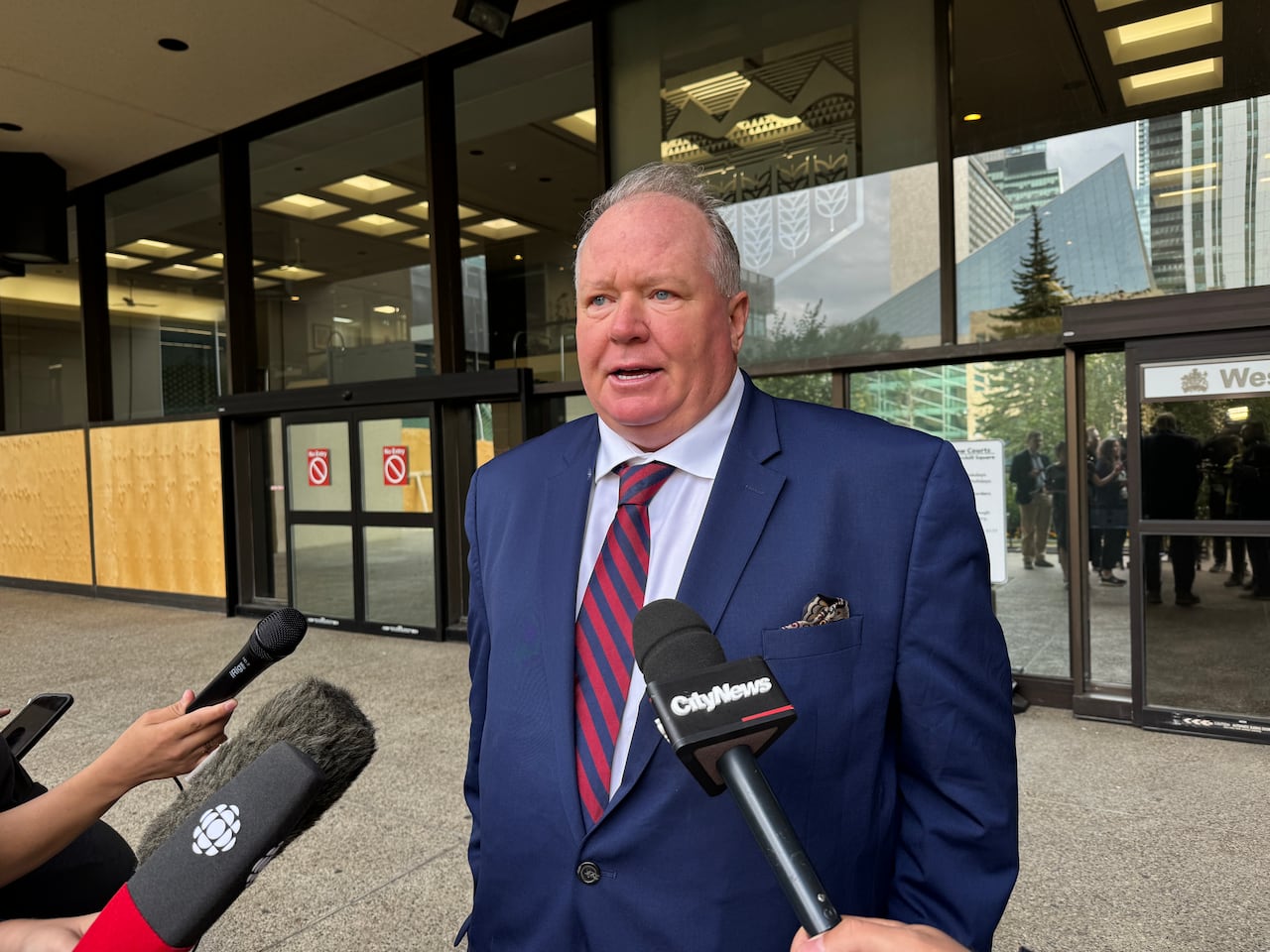 A man with grey hair, a blue suit jacket and a red striped tie stands outside the front entrance of the Edmonton court house.