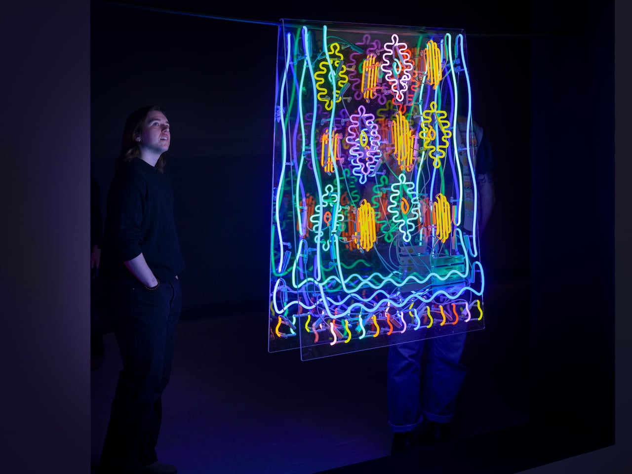 A person looks at a neon sculpture in the shape of a rug. The multicoloured artwork glows, illuminating a dark room.
