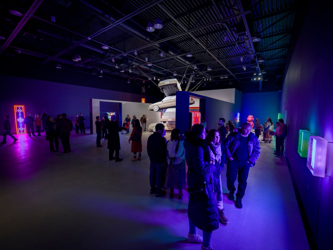 A crowd of people gathers inside a darkened gallery displaying artworks made of neon, inflatables, 3D-printed plastic, video and textiles.