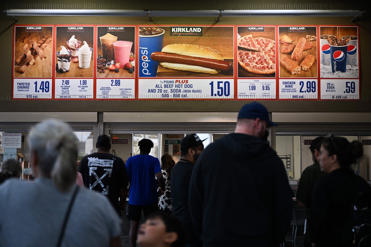 People stand in front of a food stand. Above, signs advertise hot dogs, pizza and milkshakes for sale.