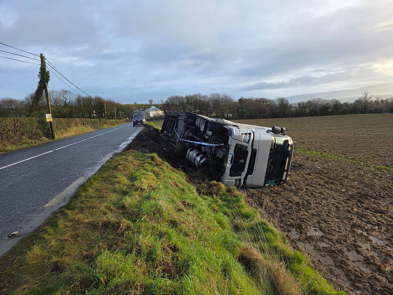 A transport truck turned on its side in a field along a narrow highway
