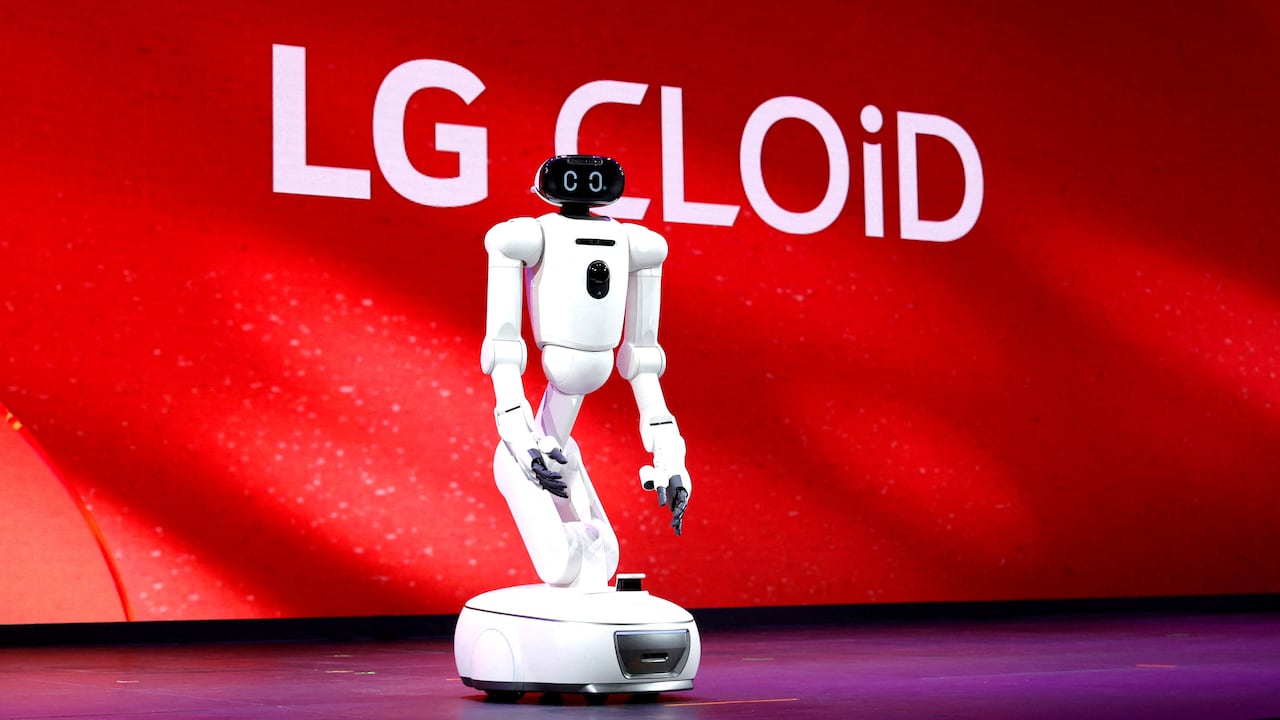 A white, humanoid robot with a sturdy round base stands on a stage with a red backdrop