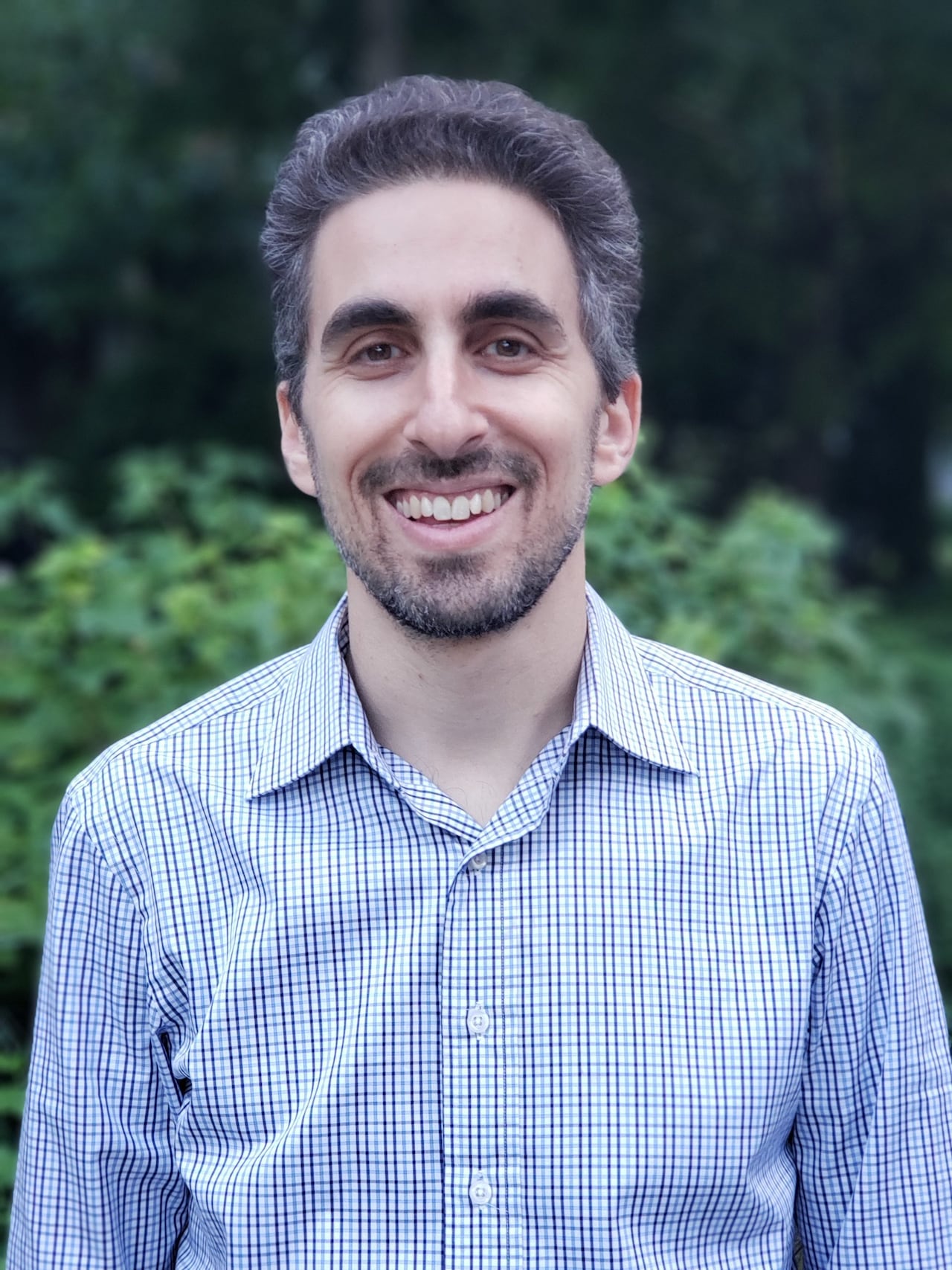 A man in a checkered shirt with a beard is smiling at the camera.