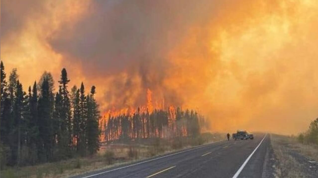 A picture of a wildfire burning in the distance as a car drives on the highway surrounded by forest.