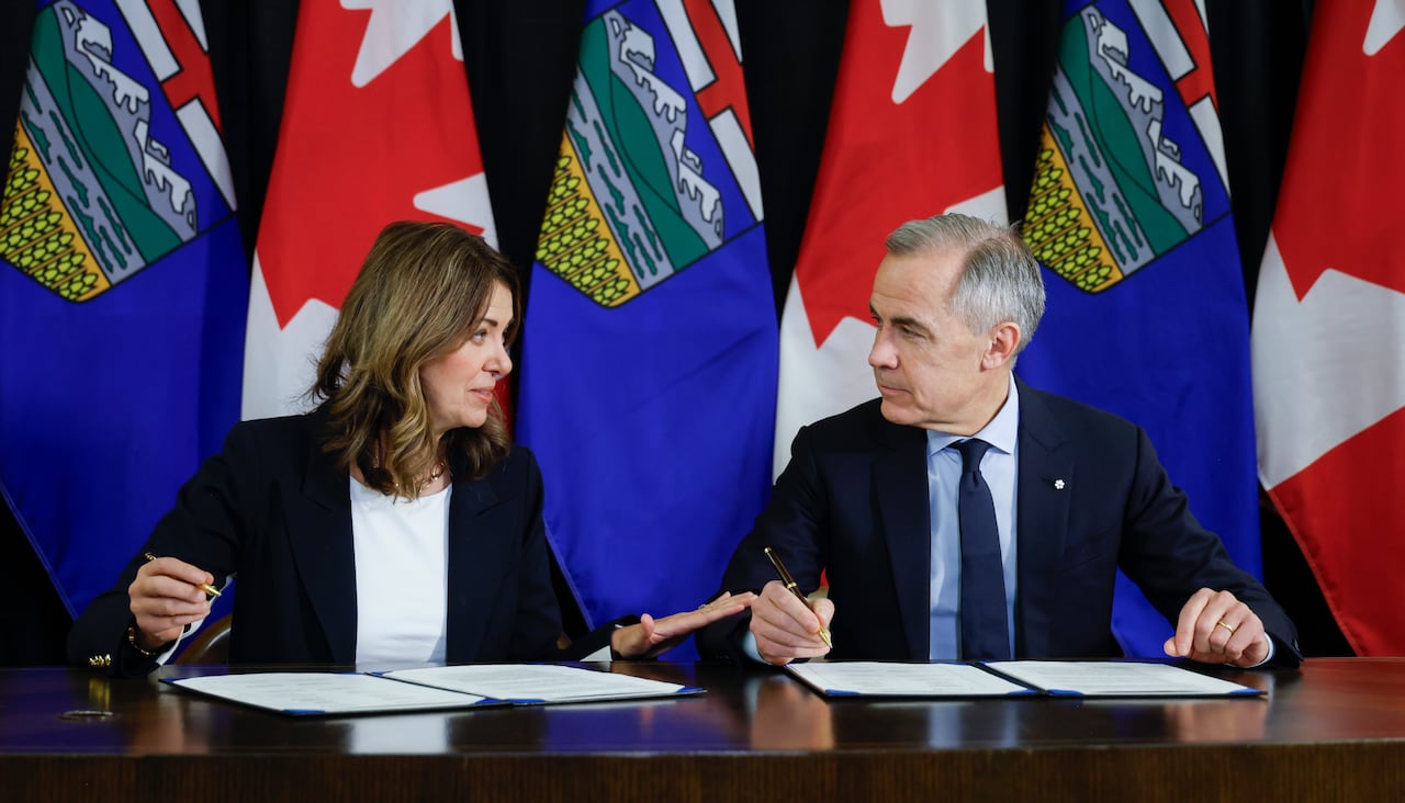With Canadian and Alberta flags in background, a woman, left, and a man, right, both in business attire are talking at a table, pen and paper in hand.